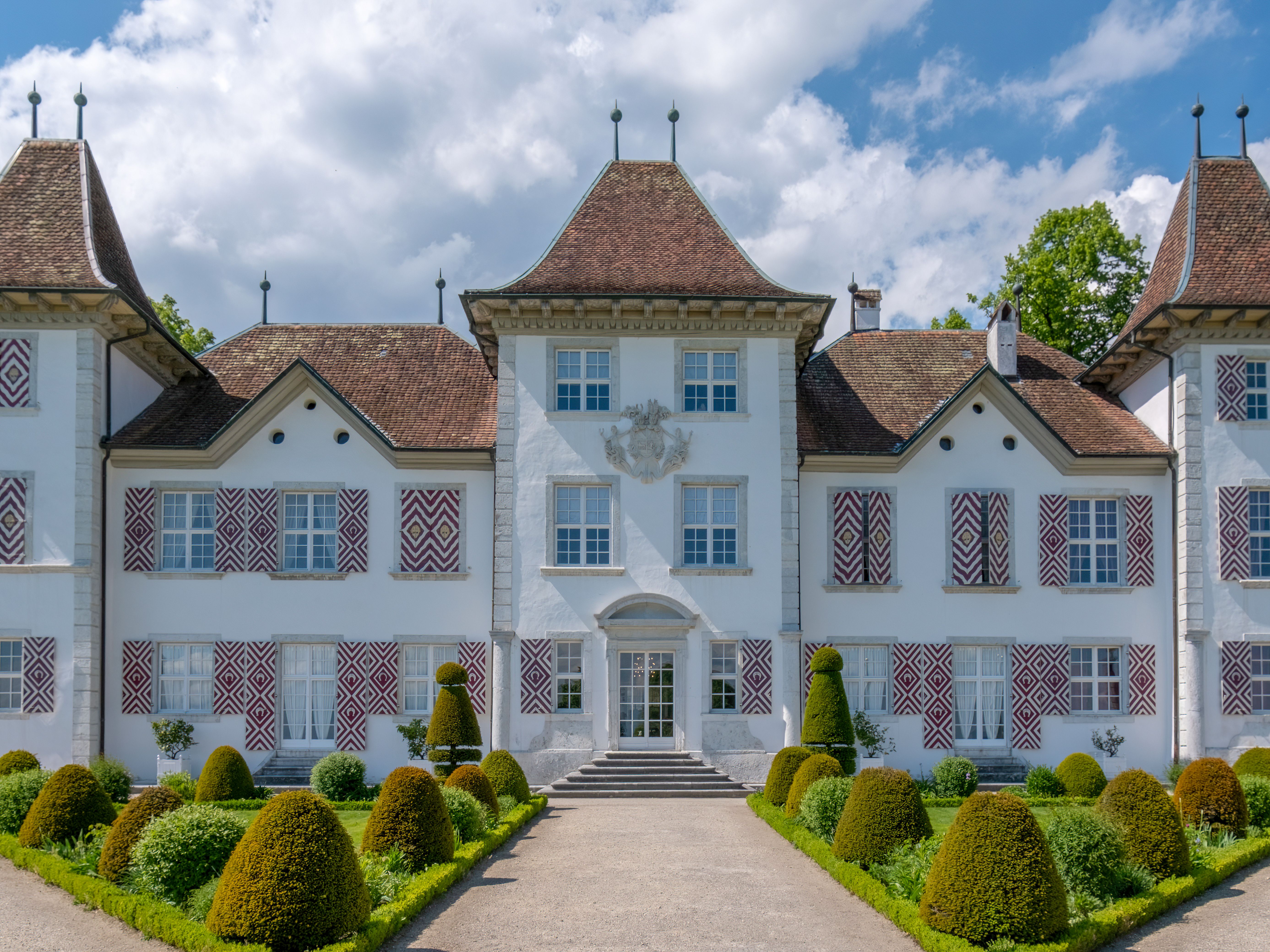 Belle demeure blanche à toitures en tuiles, entourée de jardins taillés et d’allées symétriques.